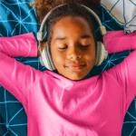 Young African-American girl in a pink shirt with headphones laying on her bed listening to music.