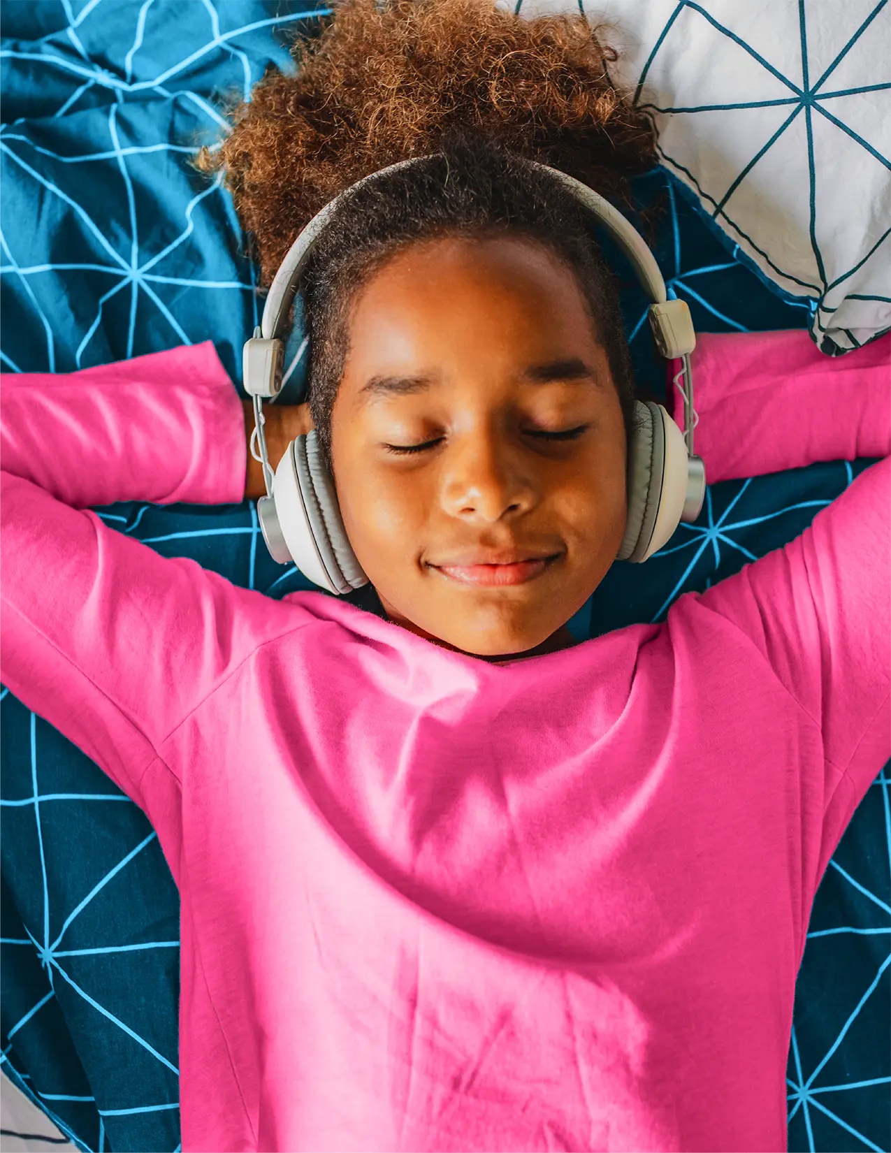 Young African-American girl in a pink shirt with headphones laying on her bed listening to music.