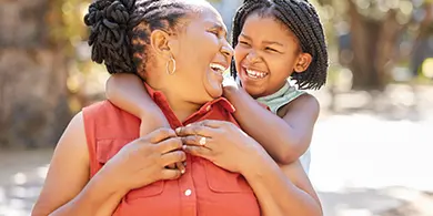 African-American mom being hugged from behind by her toddler daughter and both are laughing.