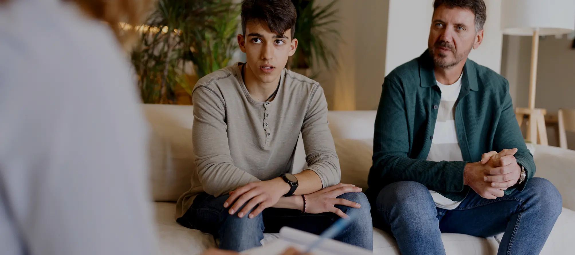 A father and son sitting on a couch talking to another person.